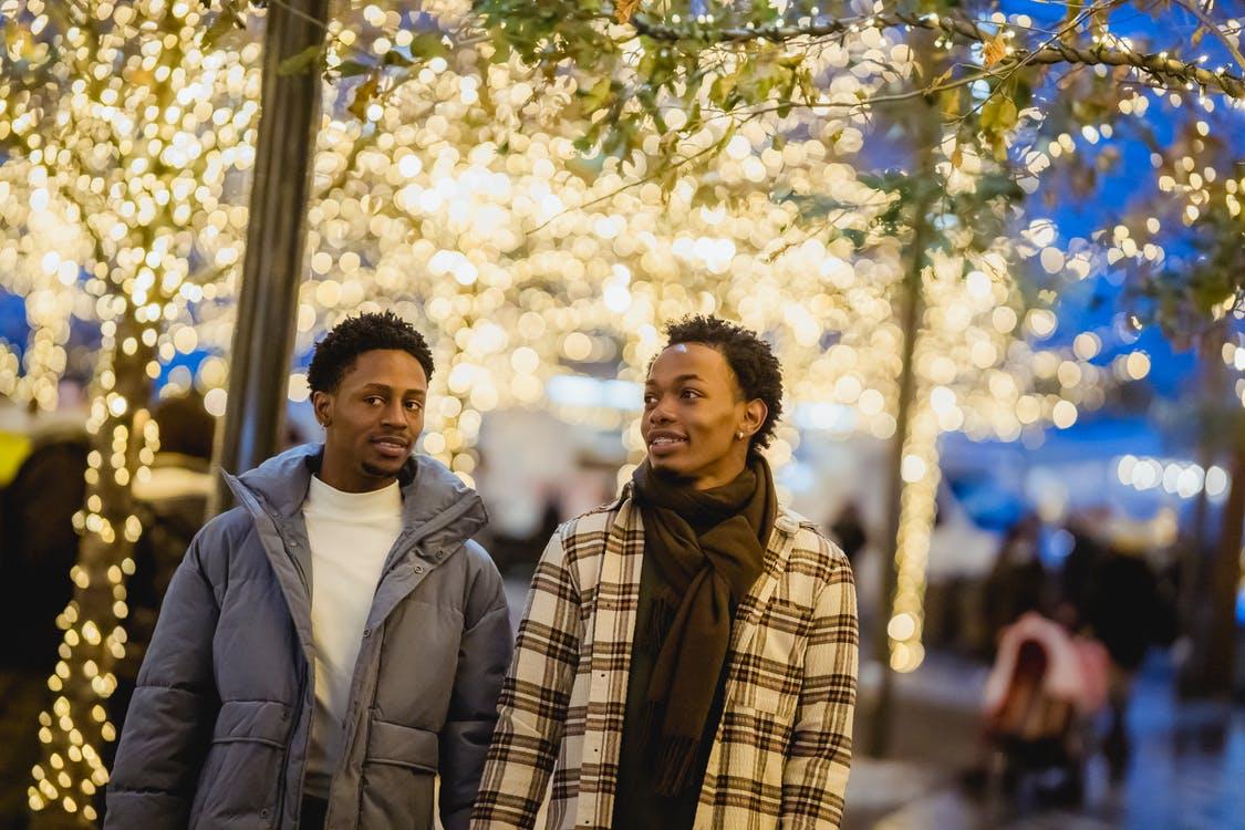 Positive African American gays strolling on street near trees with illuminated garlands at evening time and looking away on blurred background
